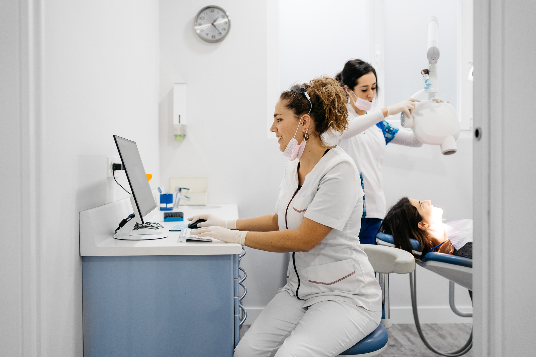 A dental professional works on a computer while another prepares an X-ray machine for a patient lying in a dental chair in a clinic.